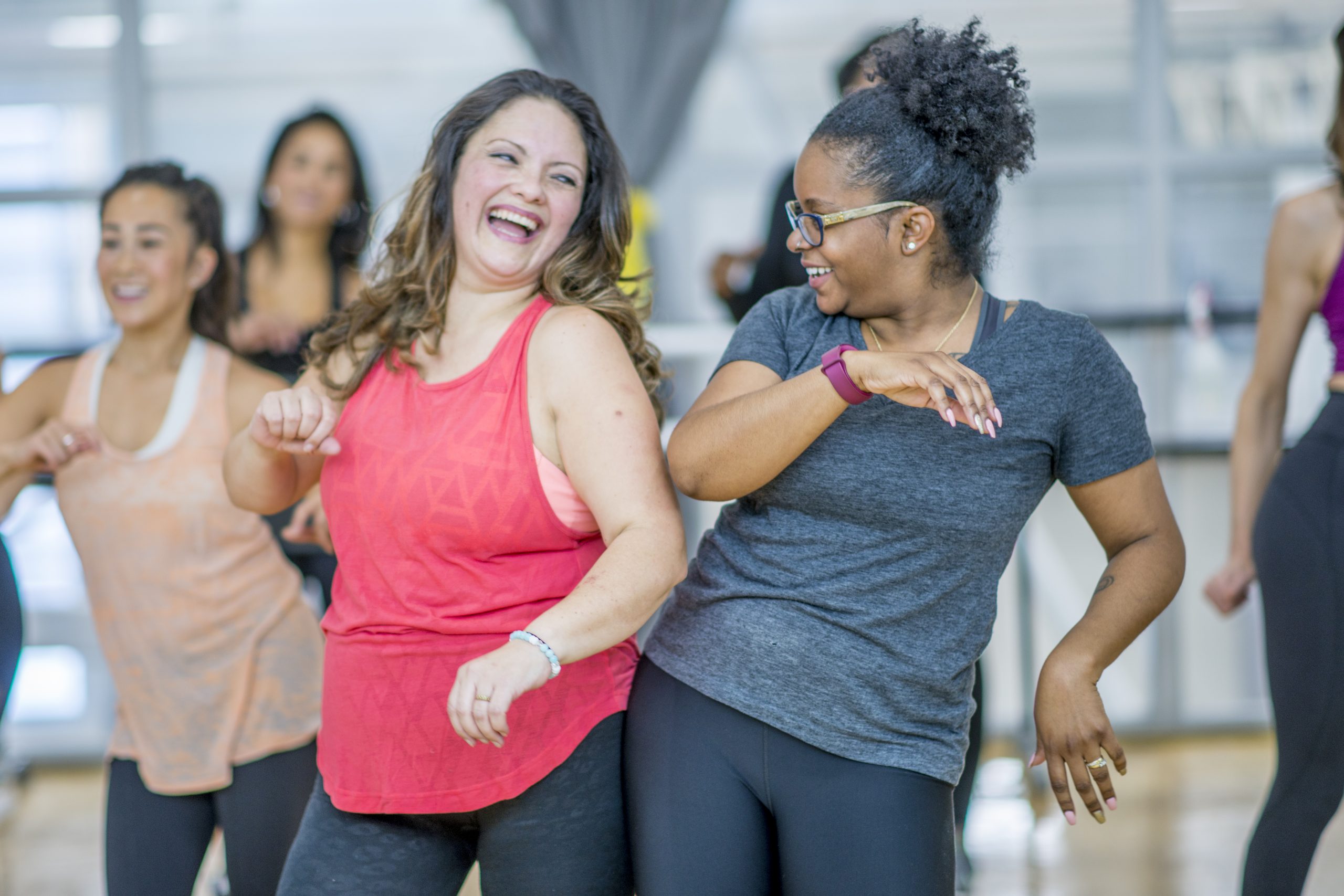 A group of young multi-ethnic women are participating in a Zumba class.  They are all wearing their active wear, smiling and having fun.  The two women in front are bumping hips and sharing a laugh.